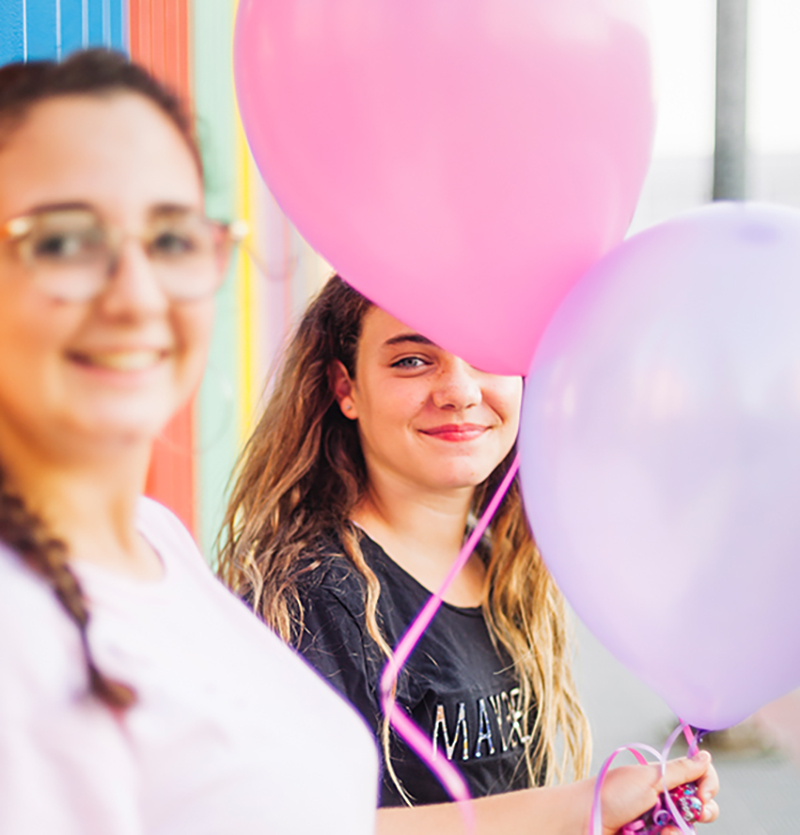 Es sind zwei junges Mädchen zu sehen. Die eine unscharf im Vordergrund und eine im Hintergrund scharf. Sie scheinen auf einer Jugendweihe oder Geburtstag zu sein.
